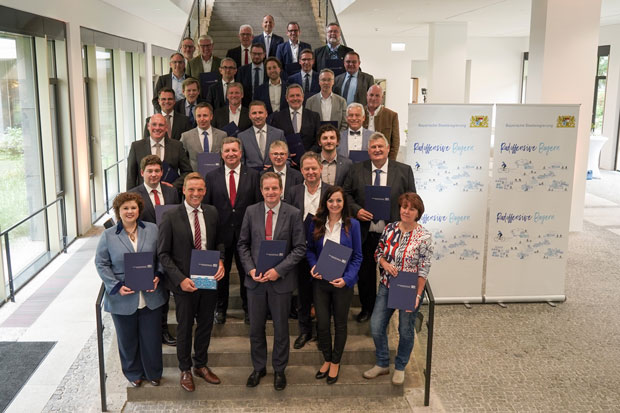 Gruppenfoto mit Verkehrsminister Christian Bernreiter und den Kommunalvertreterinnen und -vertretern für 27 Fahrrad-Projekte der Radoffensive "Klimaland Bayern". Sie stehen auf einer Treppe im Foyer des Bauministeriums . Die Kommunalvertreter halten jeweils eine Urkunde.